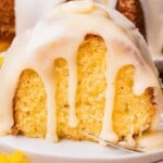 A slice of moist yellow cake with creamy glaze dripping down, served on a white plate with a yellow flower nearby.