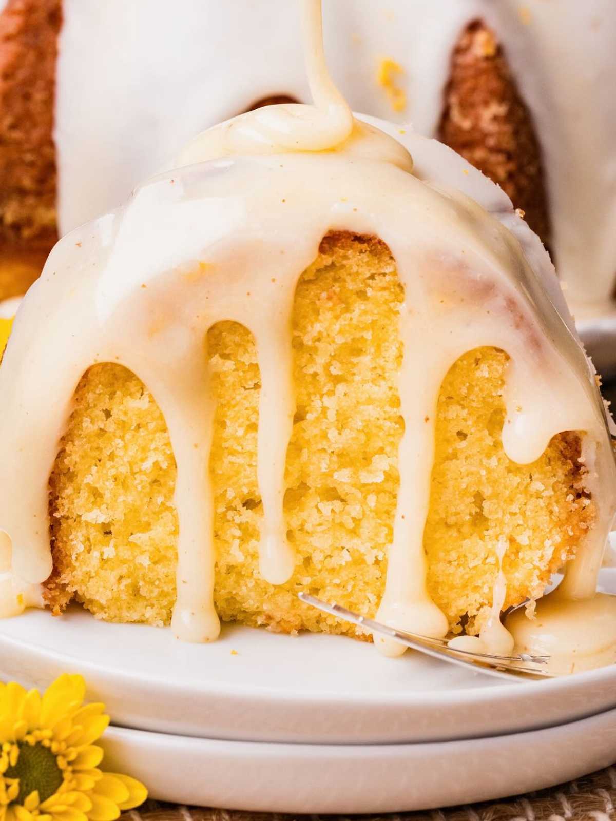 A slice of moist yellow cake with creamy glaze dripping down, served on a white plate with a yellow flower nearby.