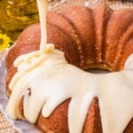 Icing being poured over a bundt cake on a plate, with a rustic background and yellow flowers nearby.