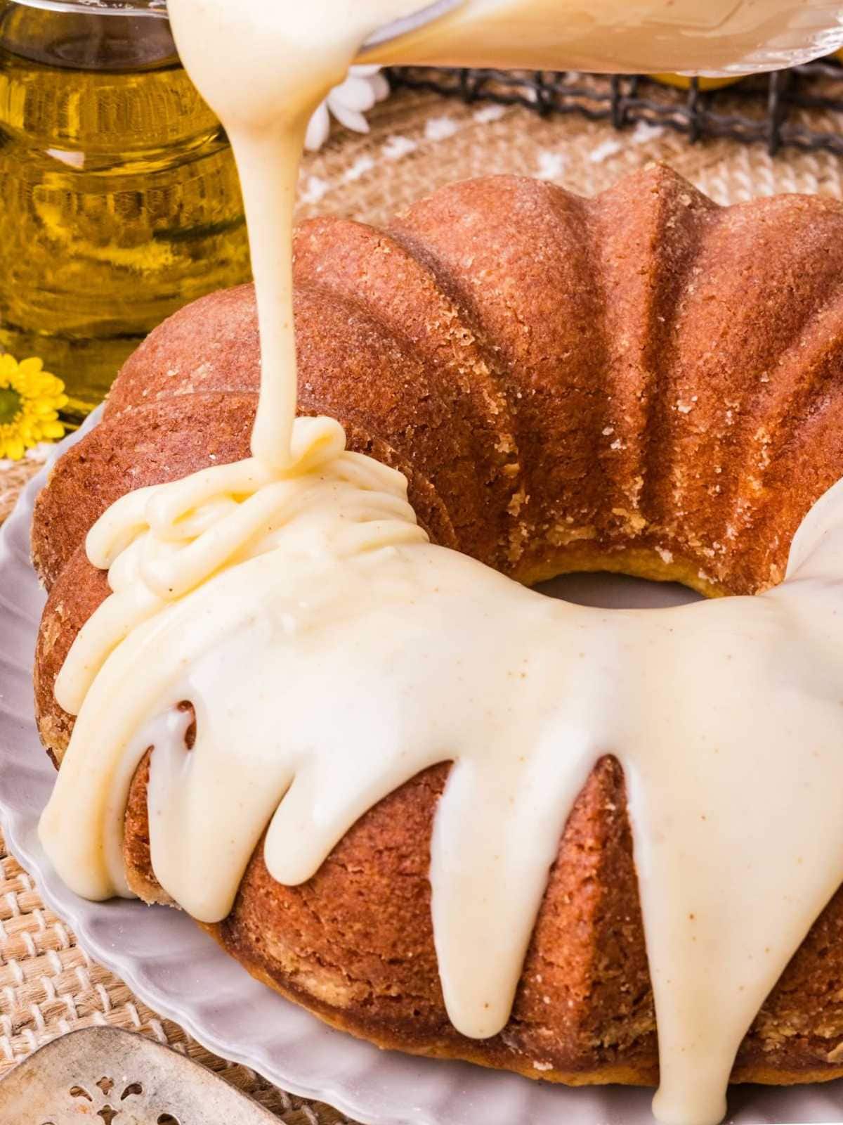 Icing being poured over a bundt cake on a plate, with a rustic background and yellow flowers nearby.
