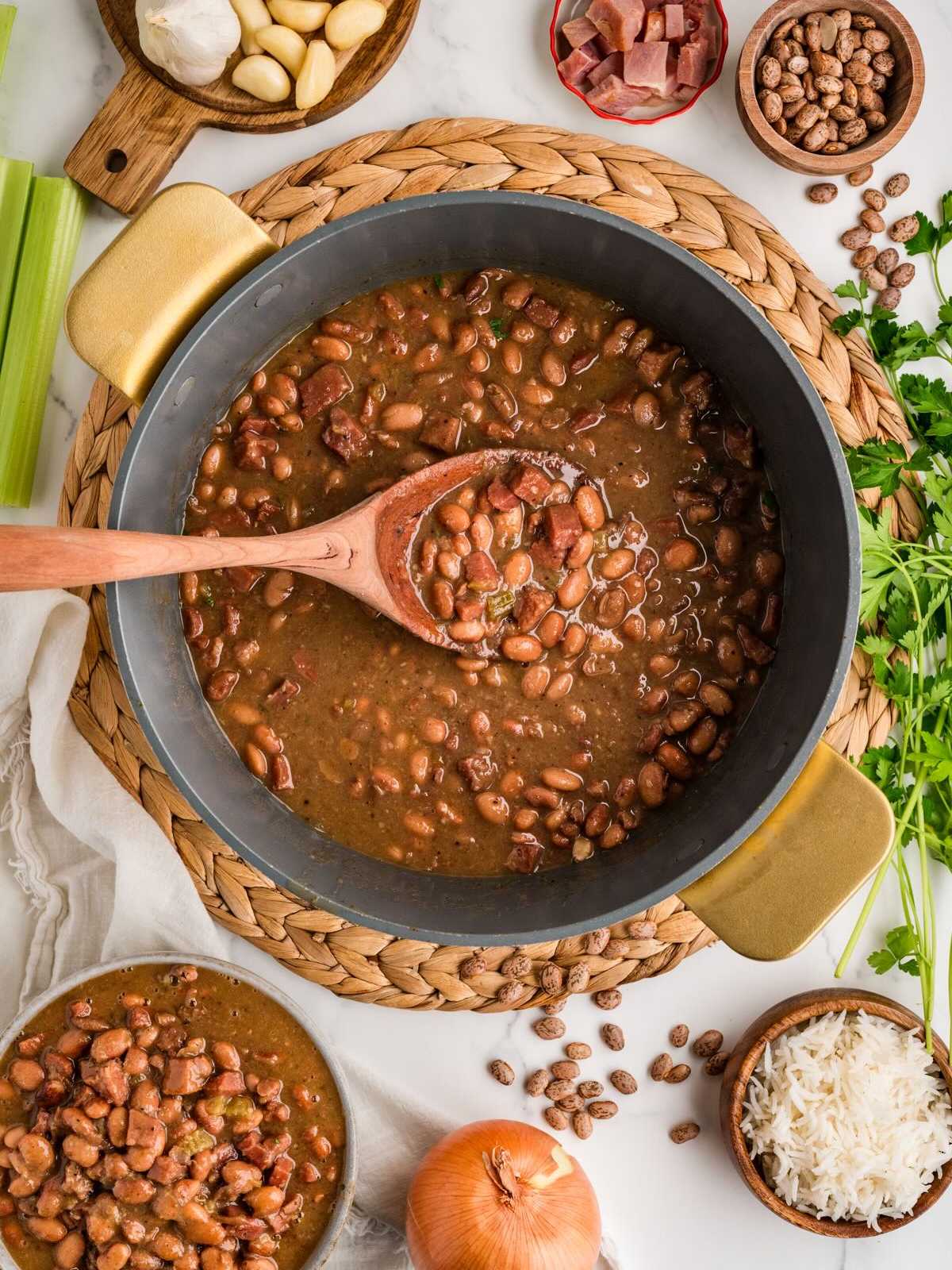A pot of cooked beans with a wooden spoon, surrounded by rice, celery, onion, garlic, herbs, and seasonings.