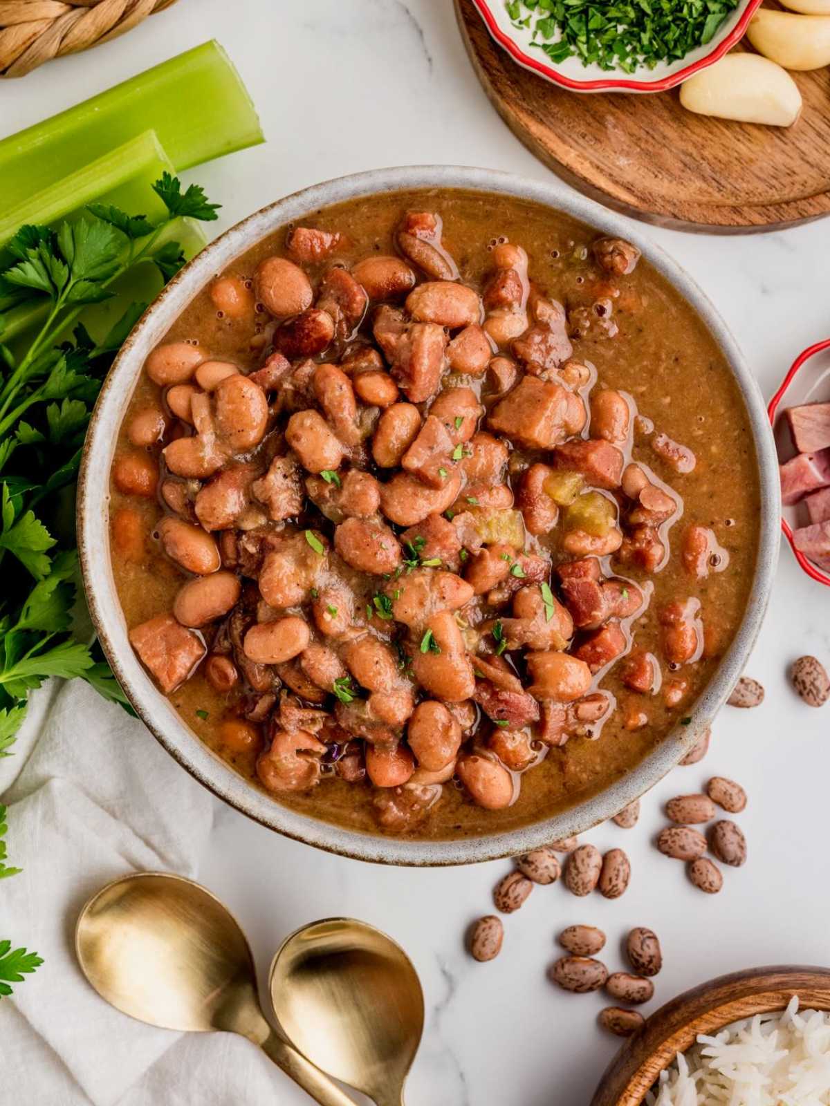 A bowl of cooked pinto beans in broth, surrounded by celery, herbs, garlic, and gold spoons on a table.