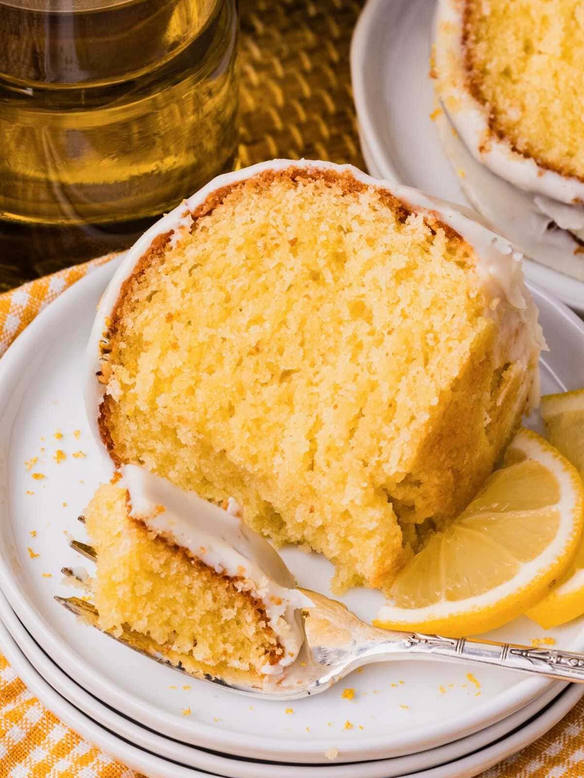 A slice of lemon cake with icing on a plate, served with lemon slices and a fork.