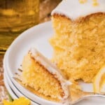 A slice of lemon cake with white icing on a plate, with a fork holding a piece and a lemon slice beside it.