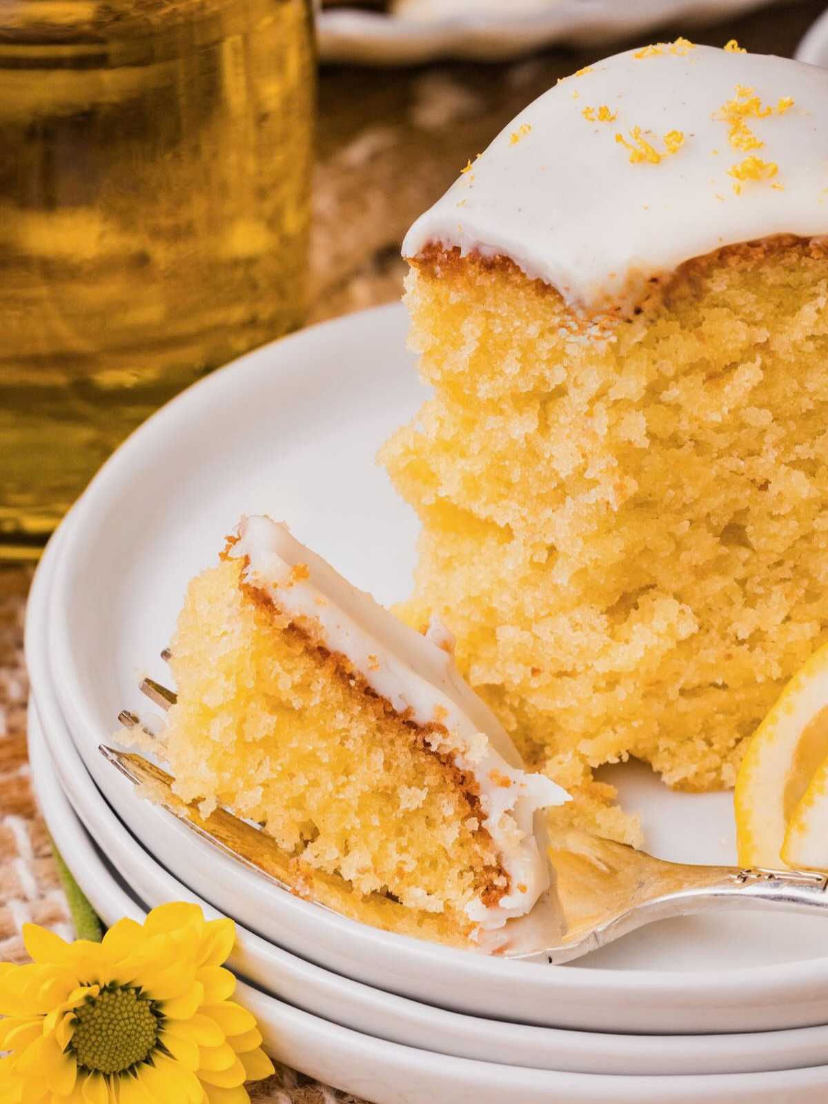 A slice of lemon cake with white icing on a plate, with a fork holding a piece and a lemon slice beside it.