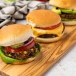 Three veggie burgers on a wooden board, topped with lettuce, tomato, onion, pickles, and cheese.