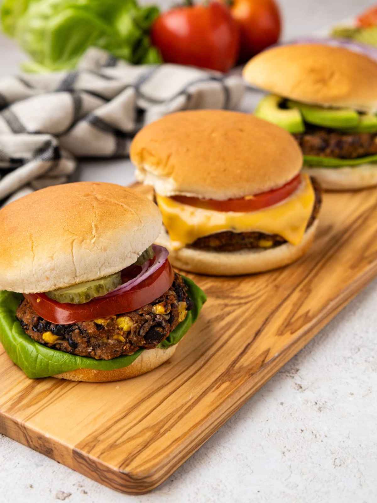 Three veggie burgers on a wooden board, topped with lettuce, tomato, onion, pickles, and cheese.