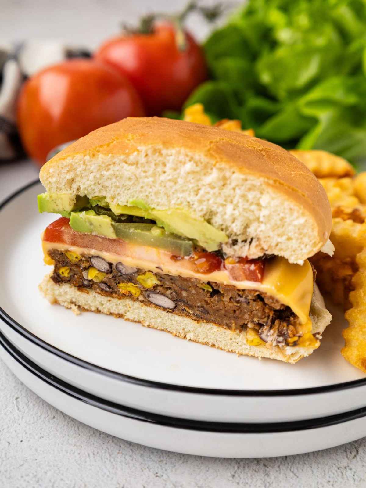 A veggie burger with cheese, avocado, tomato, and pickles, served with crinkle-cut fries on a white plate.