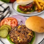 A black bean veggie burger with lettuce, pickles, tomato, and onion on a plate, with fries in the background.