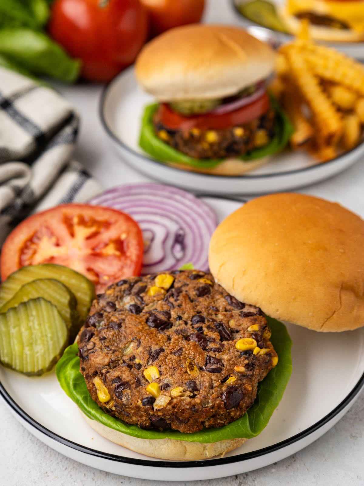A black bean veggie burger with lettuce, pickles, tomato, and onion on a plate, with fries in the background.
