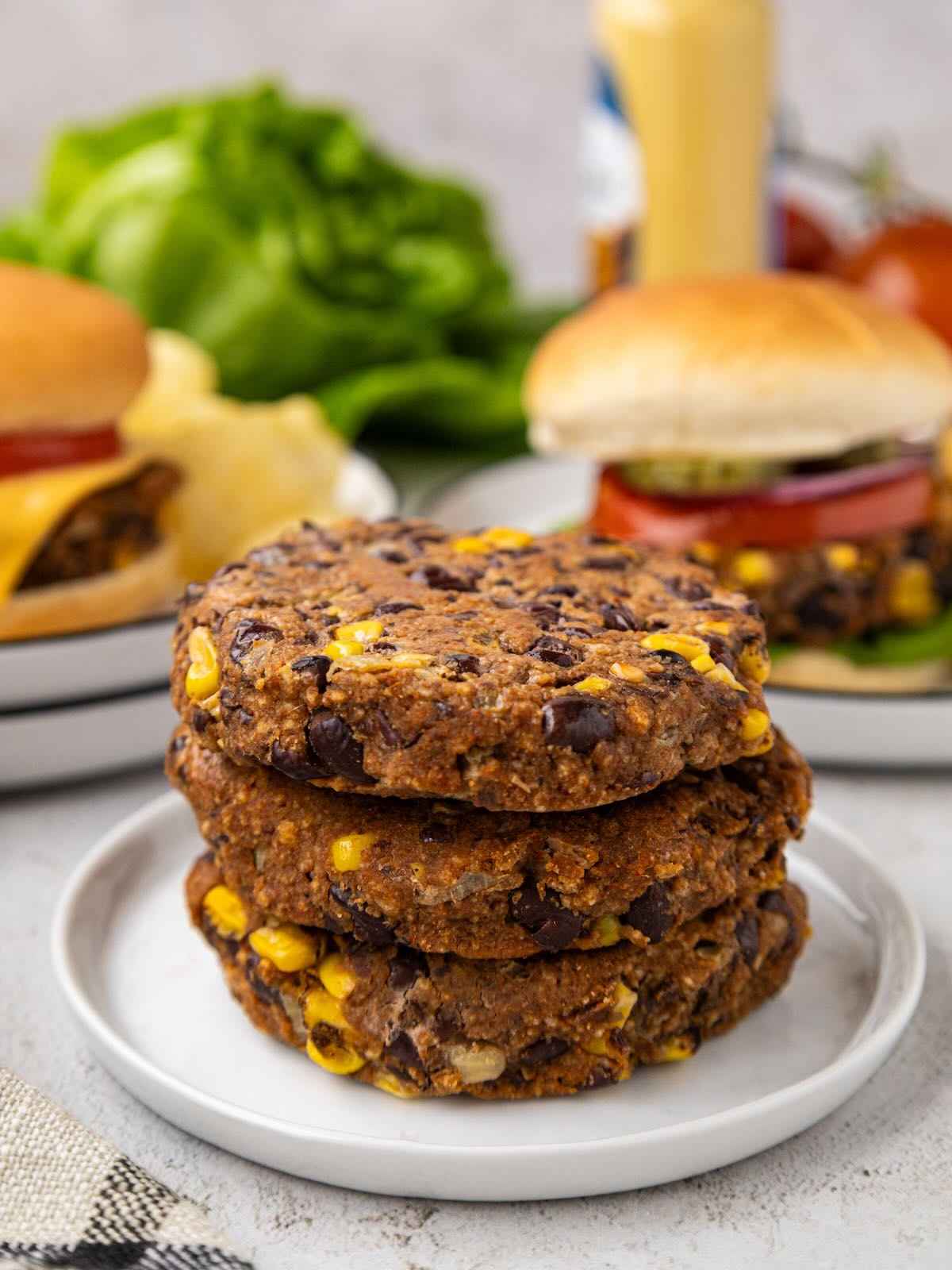Three black bean veggie burger patties stacked on a plate, with veggie burgers and lettuce in the background.