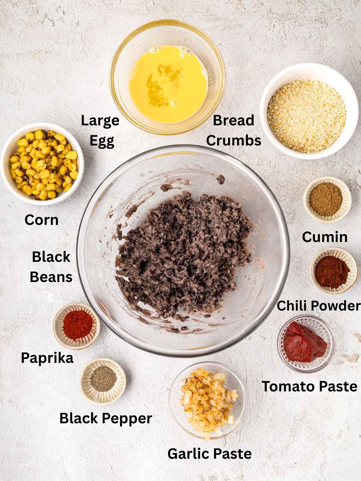 A bowl of mashed black beans surrounded by small bowls of egg, corn, bread crumbs, spices, and other ingredients.