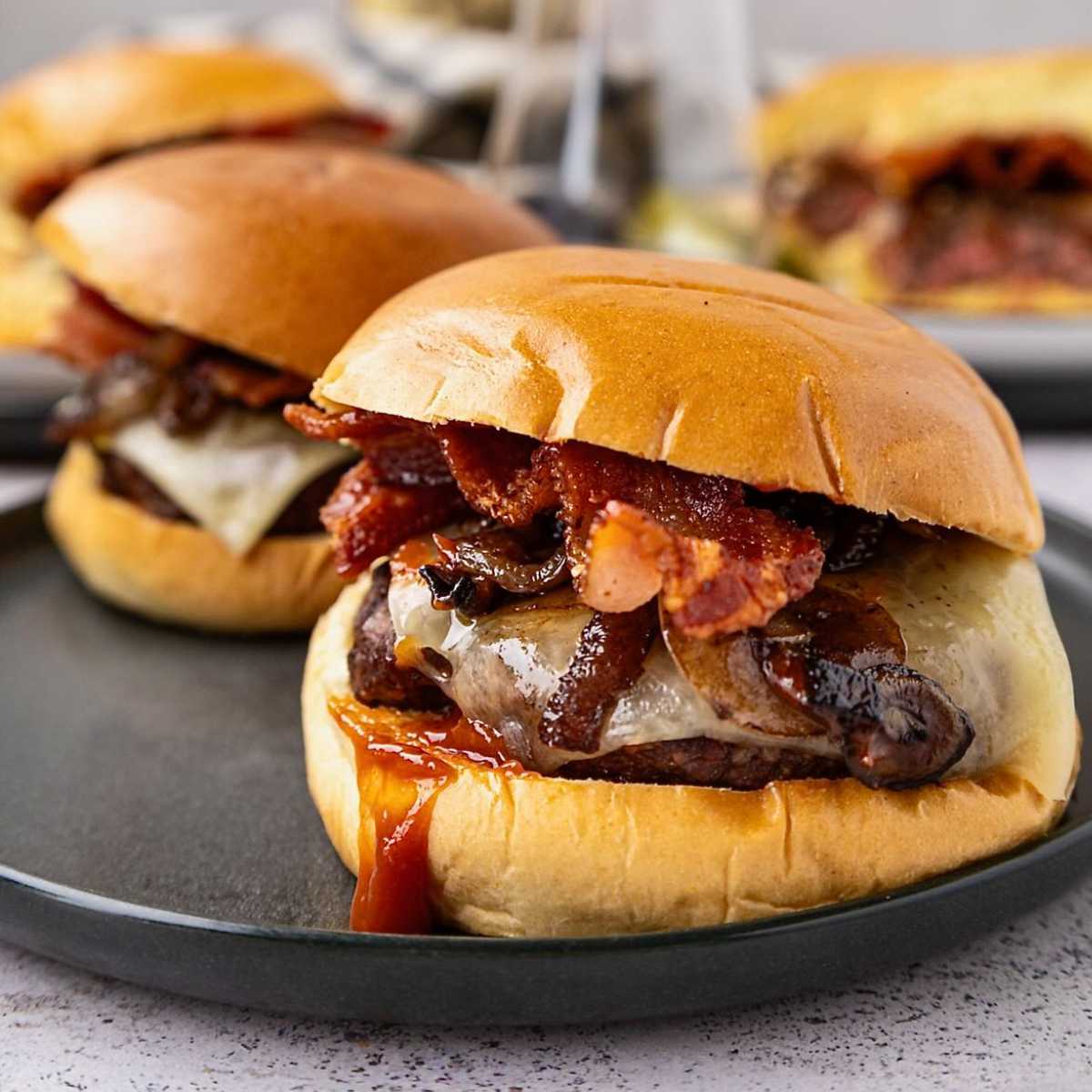 Close-up of a cheeseburger with bacon, mushrooms, and sauce on a bun, served on a dark plate.