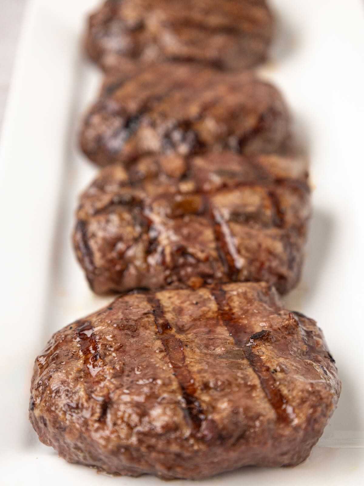Four grilled hamburger patties are arranged in a line on a white rectangular plate.