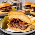 A sliced burger with bacon and cheese on a plate, served with potato chips and a pickle spear.