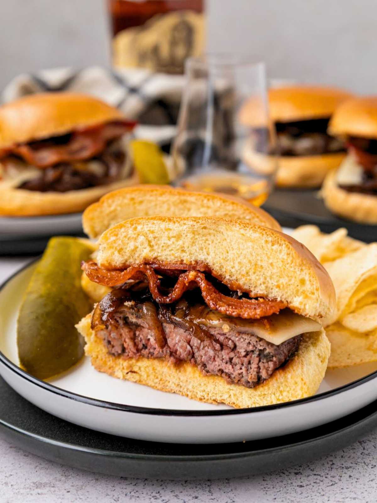 A sliced burger with bacon and cheese on a plate, served with potato chips and a pickle spear.