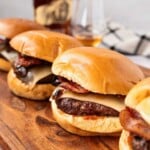 Three cheeseburgers with bacon on a wooden board, with a bottle and glasses in the background.