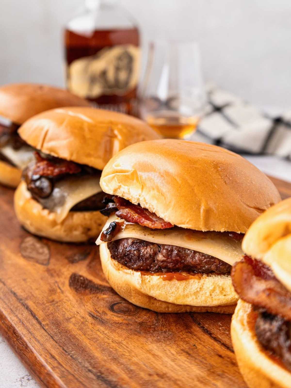 Three cheeseburgers with bacon on a wooden board, with a bottle and glasses in the background.