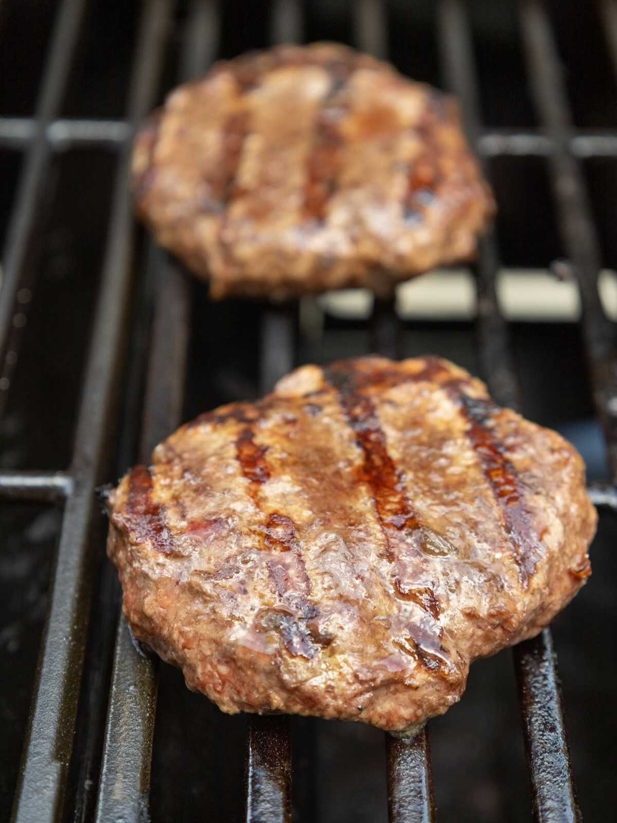 Two beef burger patties with grill marks cooking on a barbecue grill.