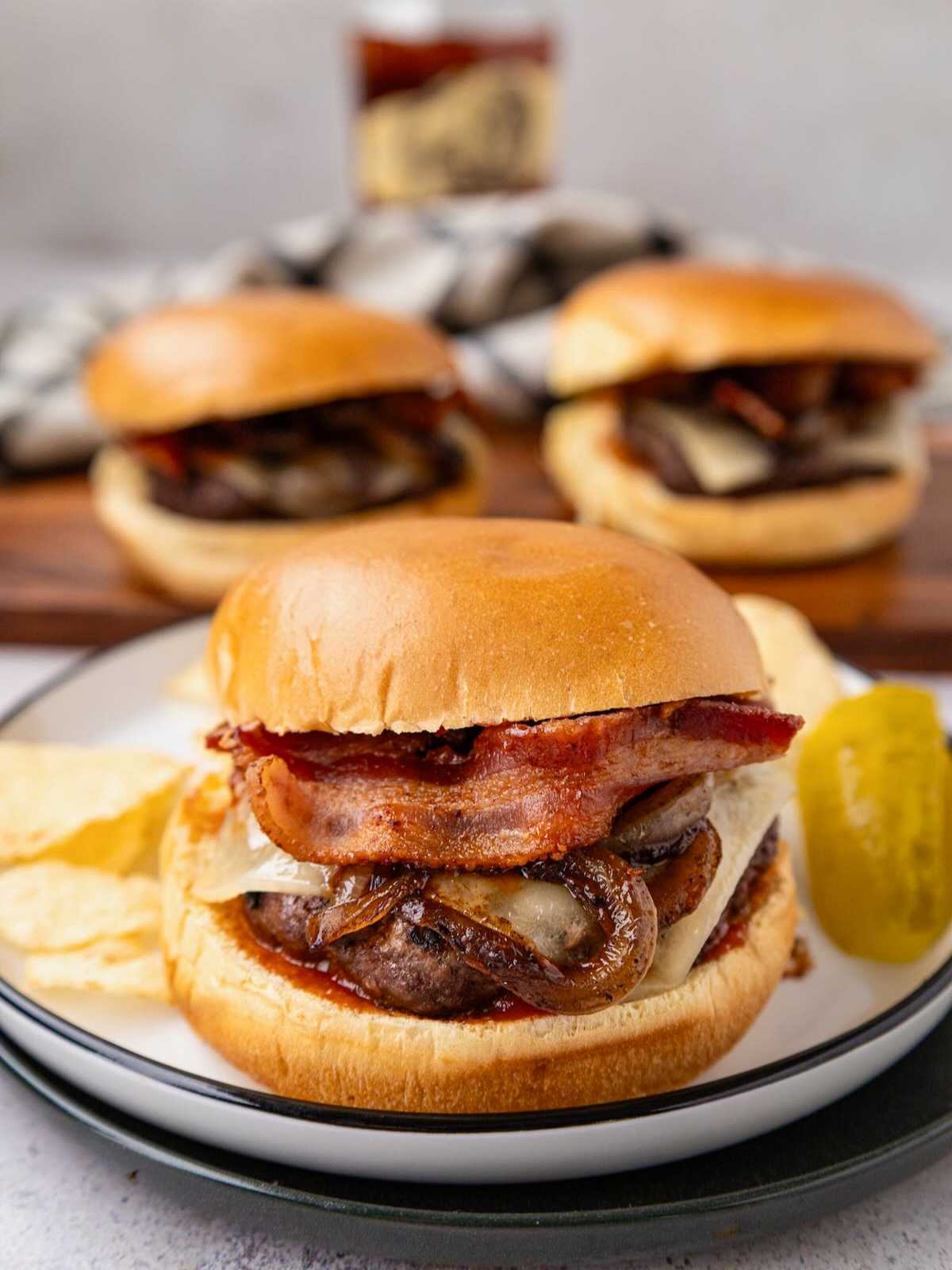 A cheeseburger with bacon, onions, chips, and a pickle on a plate; two more burgers in the background.