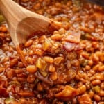 A wooden spoon scooping cowboy baked beans with bacon and vegetables from a pot.