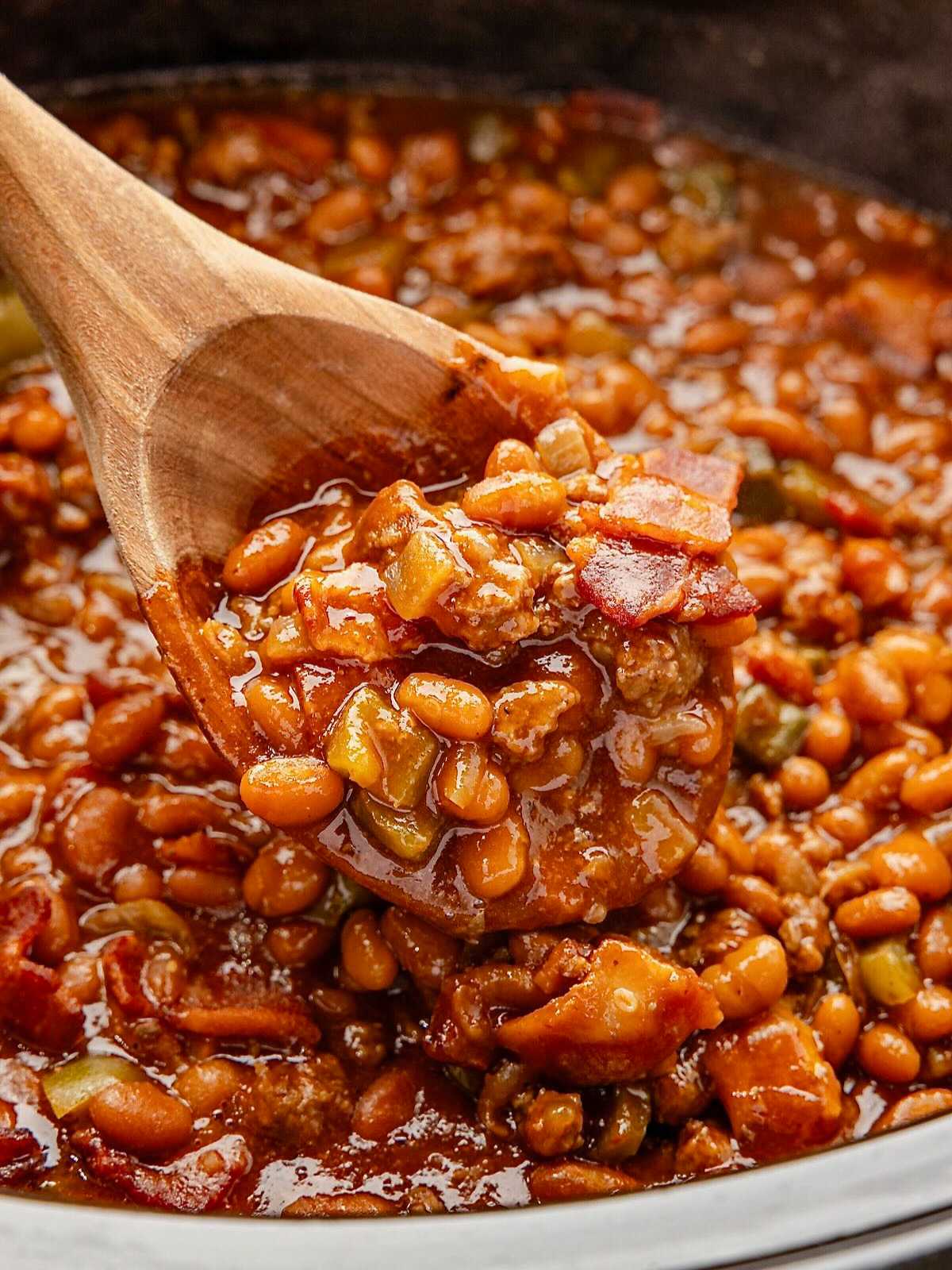 A wooden spoon scooping cowboy baked beans with bacon and vegetables from a pot.
