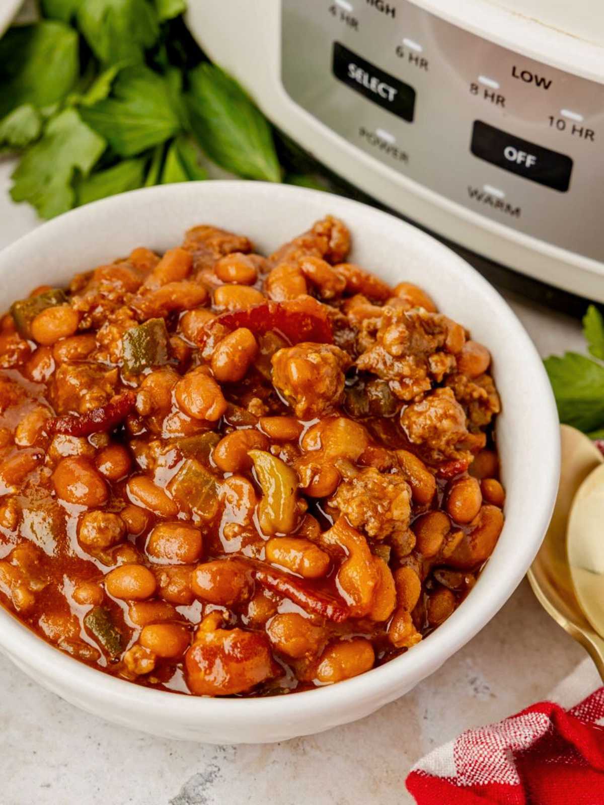 A bowl of cowboy beans and ground meat sits in front of a slow cooker, garnished with fresh parsley.