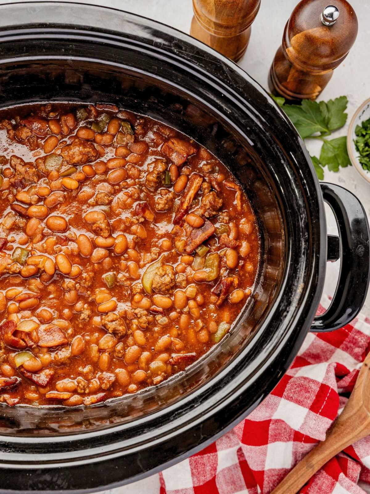 Slow-cooked baked beans with bacon and sauce in a black crockpot, next to a red checkered napkin.