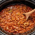 Slow cooker filled with baked beans, ground meat, and vegetables, stirred with a wooden spoon.