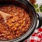 Cowboy baked beans with ground meat and bacon in a slow cooker, stirred with a wooden spoon.