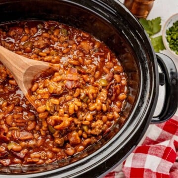 Cowboy baked beans with ground meat and bacon in a slow cooker, stirred with a wooden spoon.