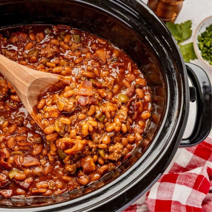 Cowboy baked beans with ground meat and bacon in a slow cooker, stirred with a wooden spoon.