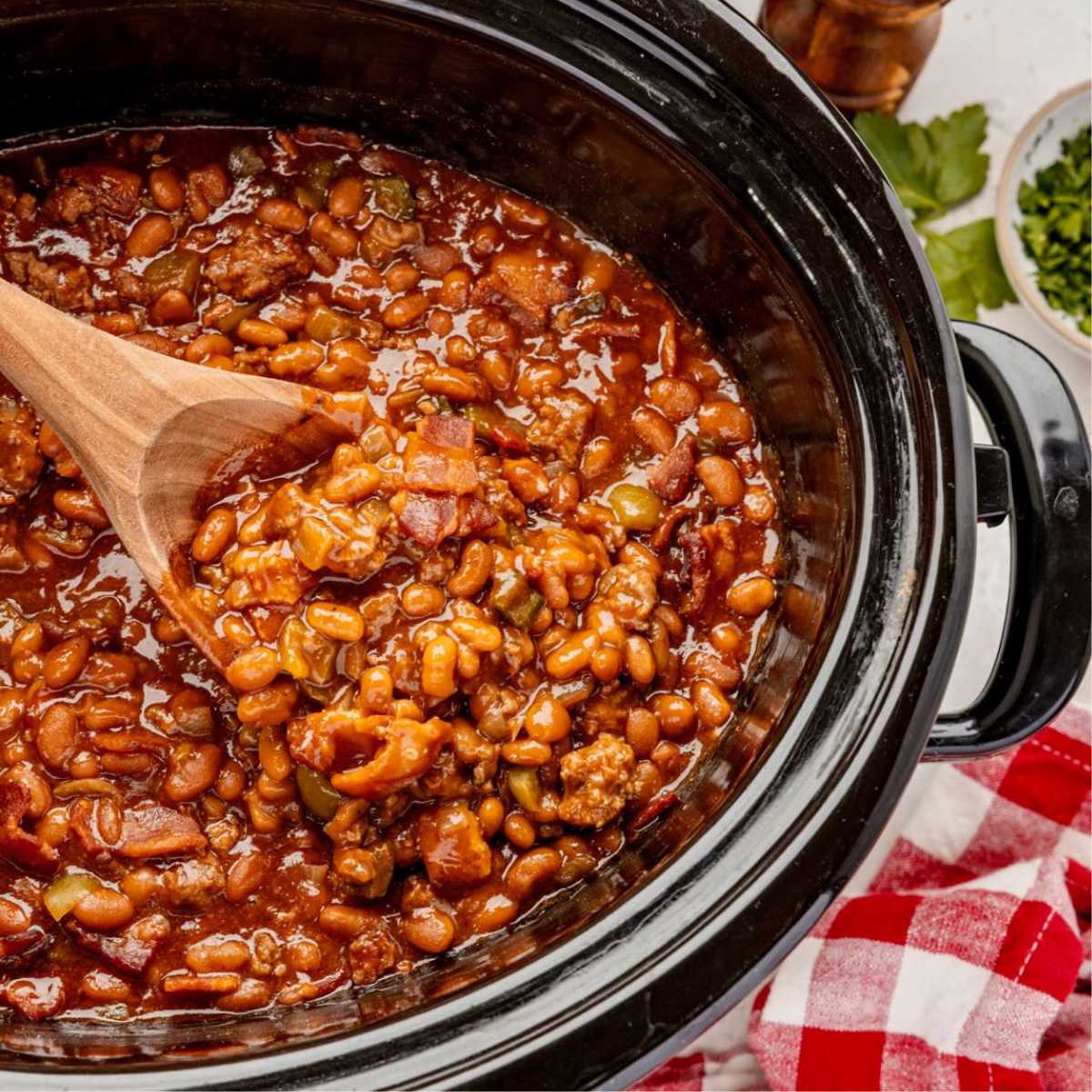 Cowboy baked beans with ground meat and bacon in a slow cooker, stirred with a wooden spoon.