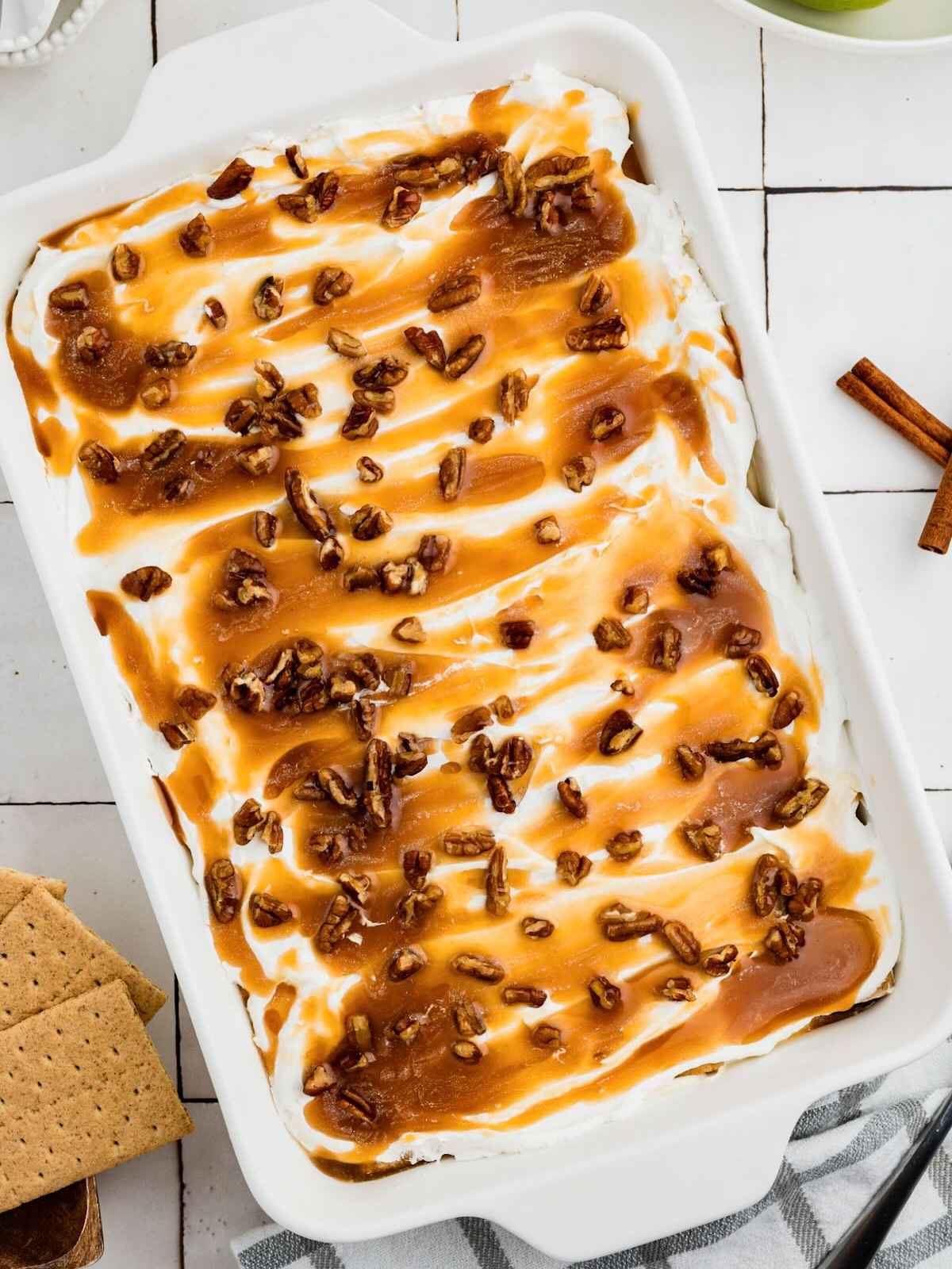 Dessert topped with whipped cream, caramel sauce, and chopped pecans in a white dish on a tiled surface.