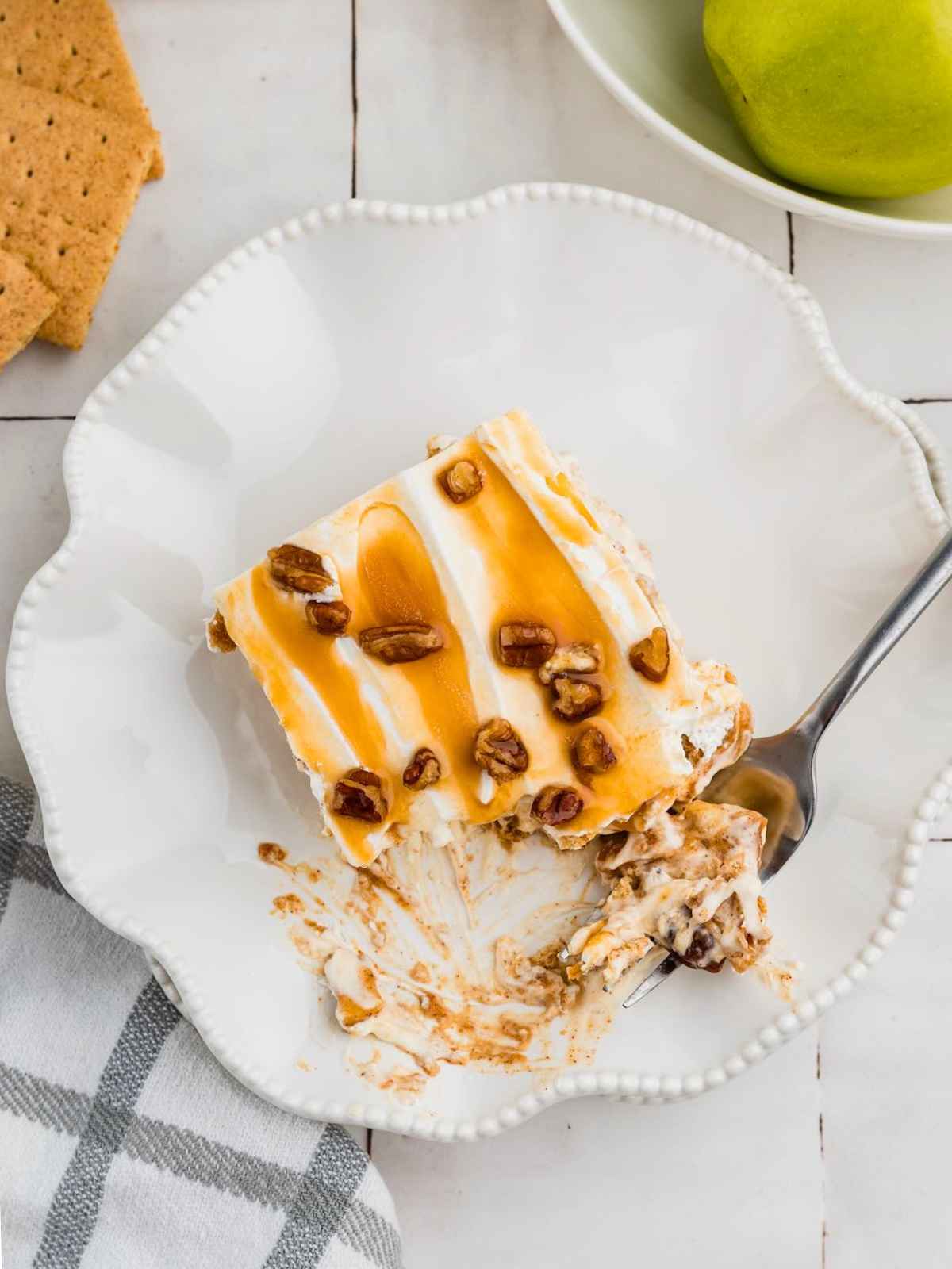 A dessert slice with caramel and pecans on a white plate, a fork, and part of an apple and napkin nearby.