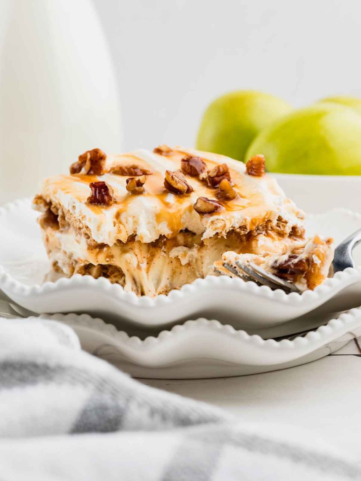 A layered caramel dessert topped with whipped cream and pecans, served on a white plate with apples in background.