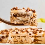 A wooden spatula holds a slice of caramel apple eclair cake with pecans above a dish of the same dessert.