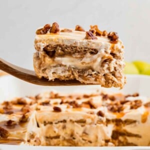 A wooden spatula holds a slice of caramel apple eclair cake with pecans above a dish of the same dessert.
