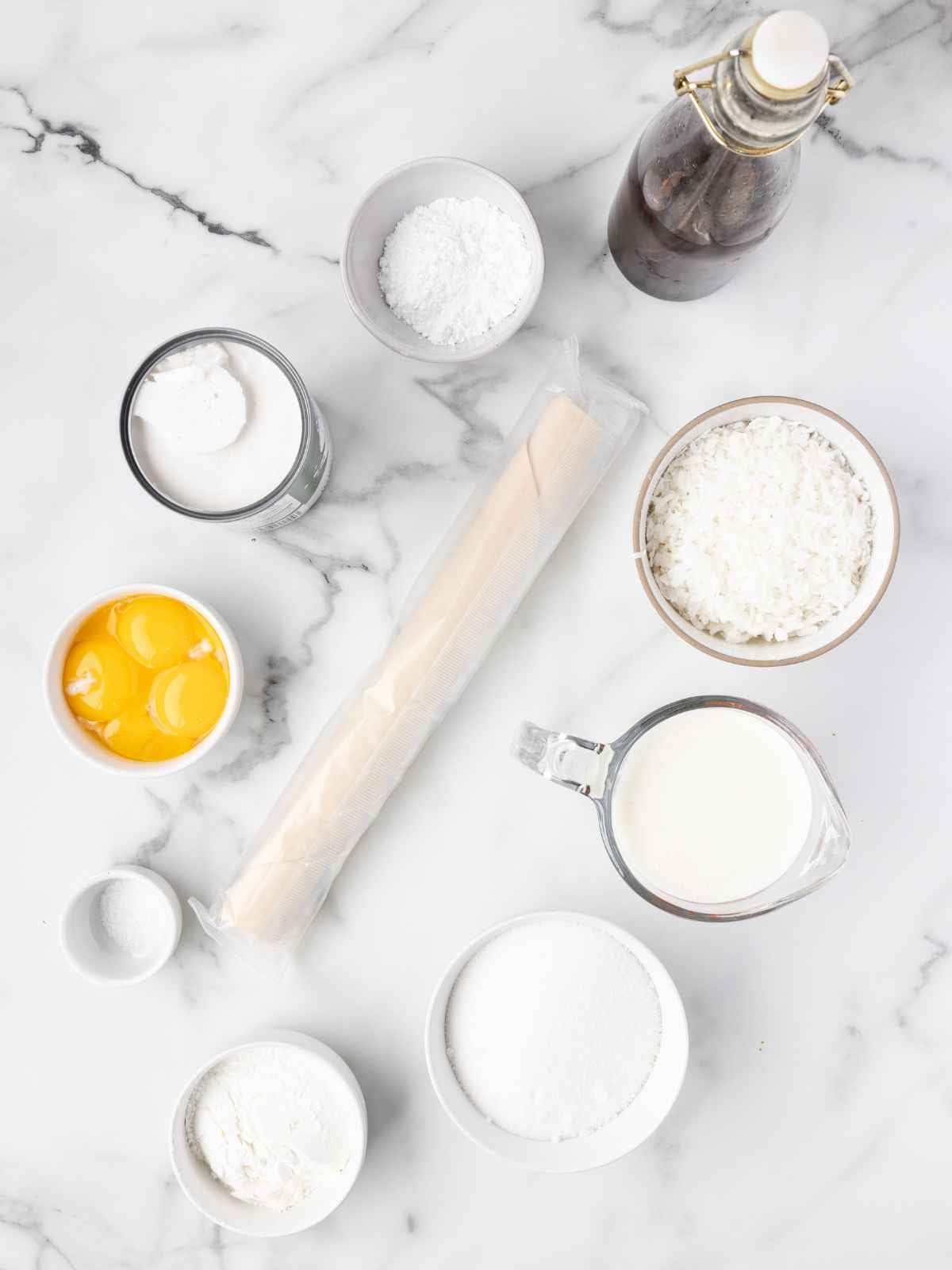 Baking ingredients on marble: eggs, cream, sugar, flour, coconut, pastry, vanilla, and powders in bowls.
