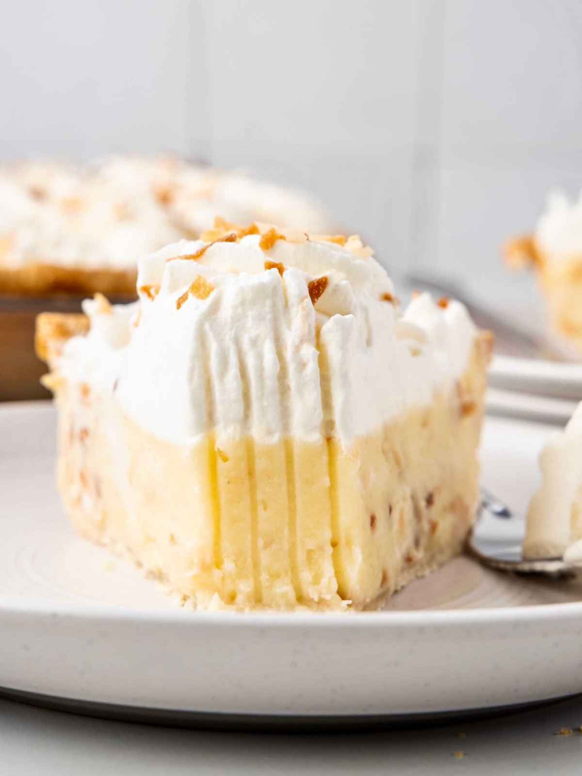 A slice of coconut cream pie with whipped topping on a white plate, bite taken from the front.