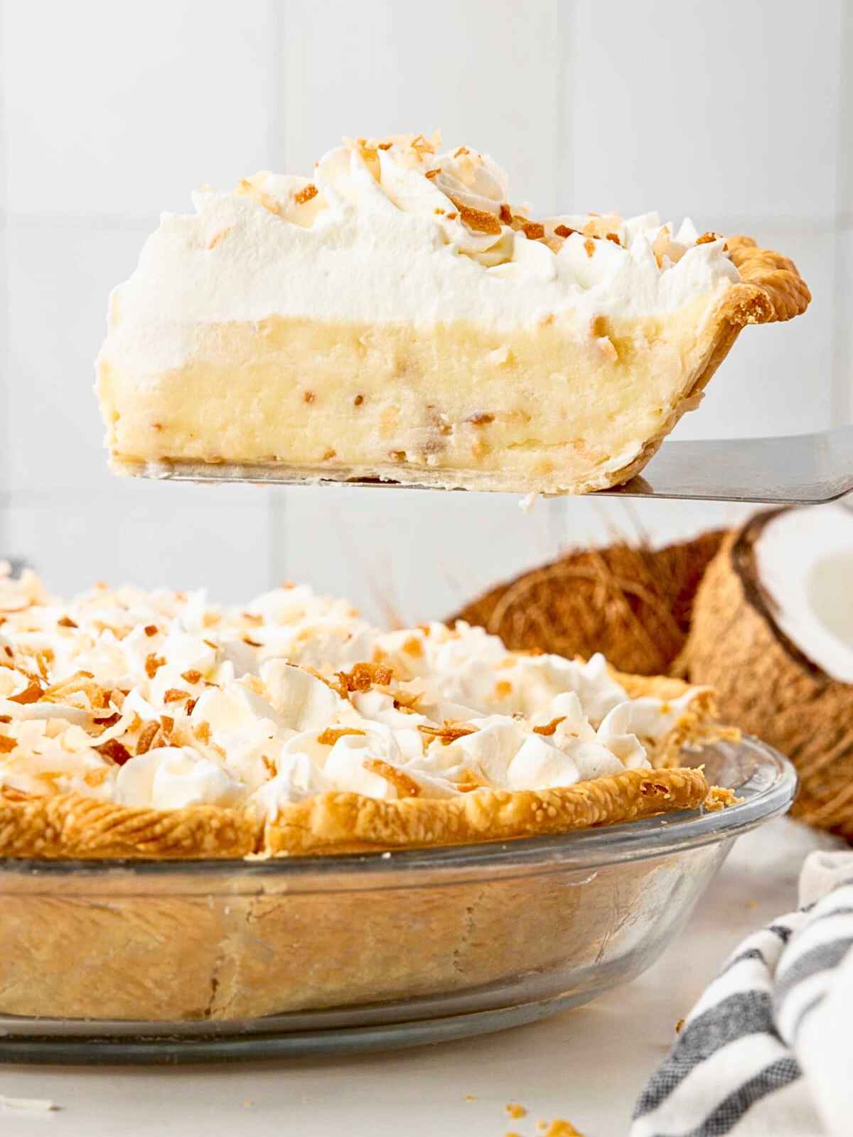 A slice of coconut cream pie with whipped topping being lifted from a whole pie in a glass dish.