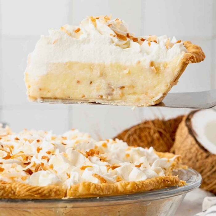 A slice of coconut cream pie with whipped topping is being lifted from a pie dish.