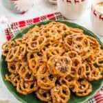 A pile of glazed, toffee pretzels on a green plate, with mugs of hot cocoa in the background.
