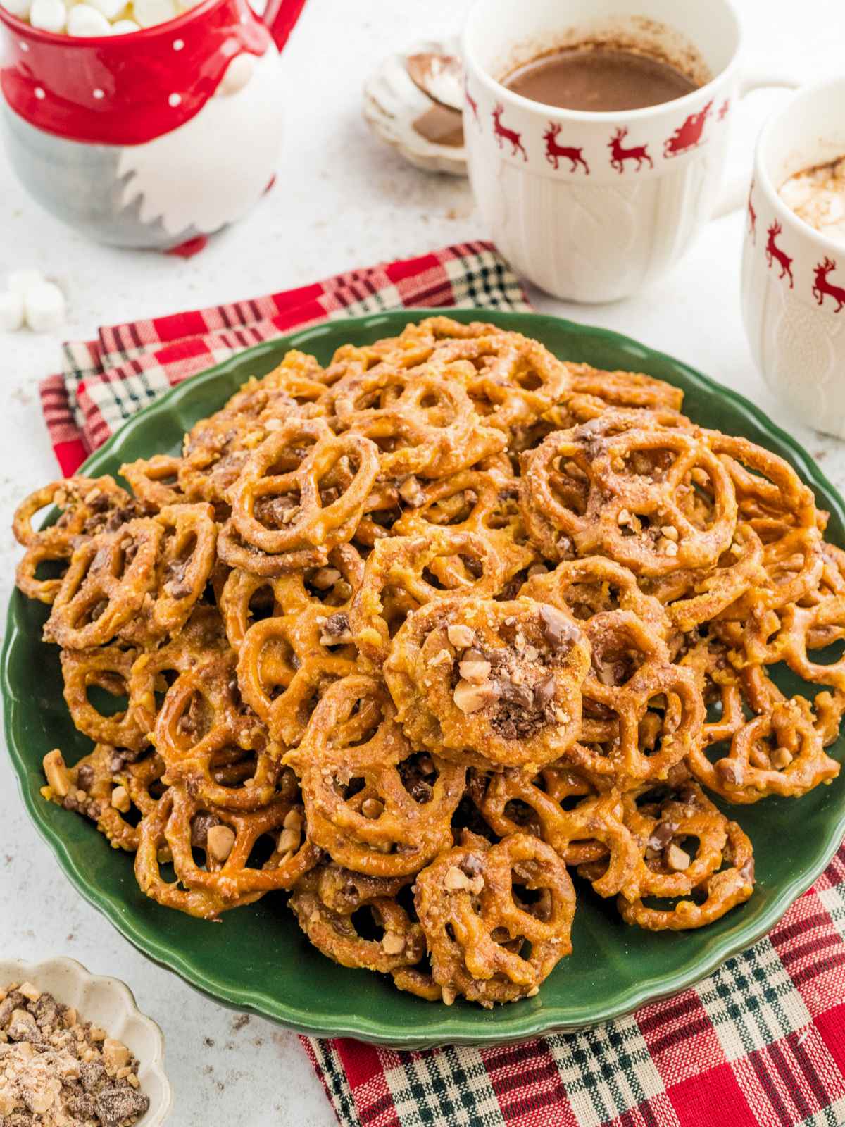 A pile of glazed, toffee pretzels on a green plate, with mugs of hot cocoa in the background.