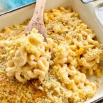A wooden spoon lifting a scoop of baked macaroni and cheese topped with breadcrumbs.