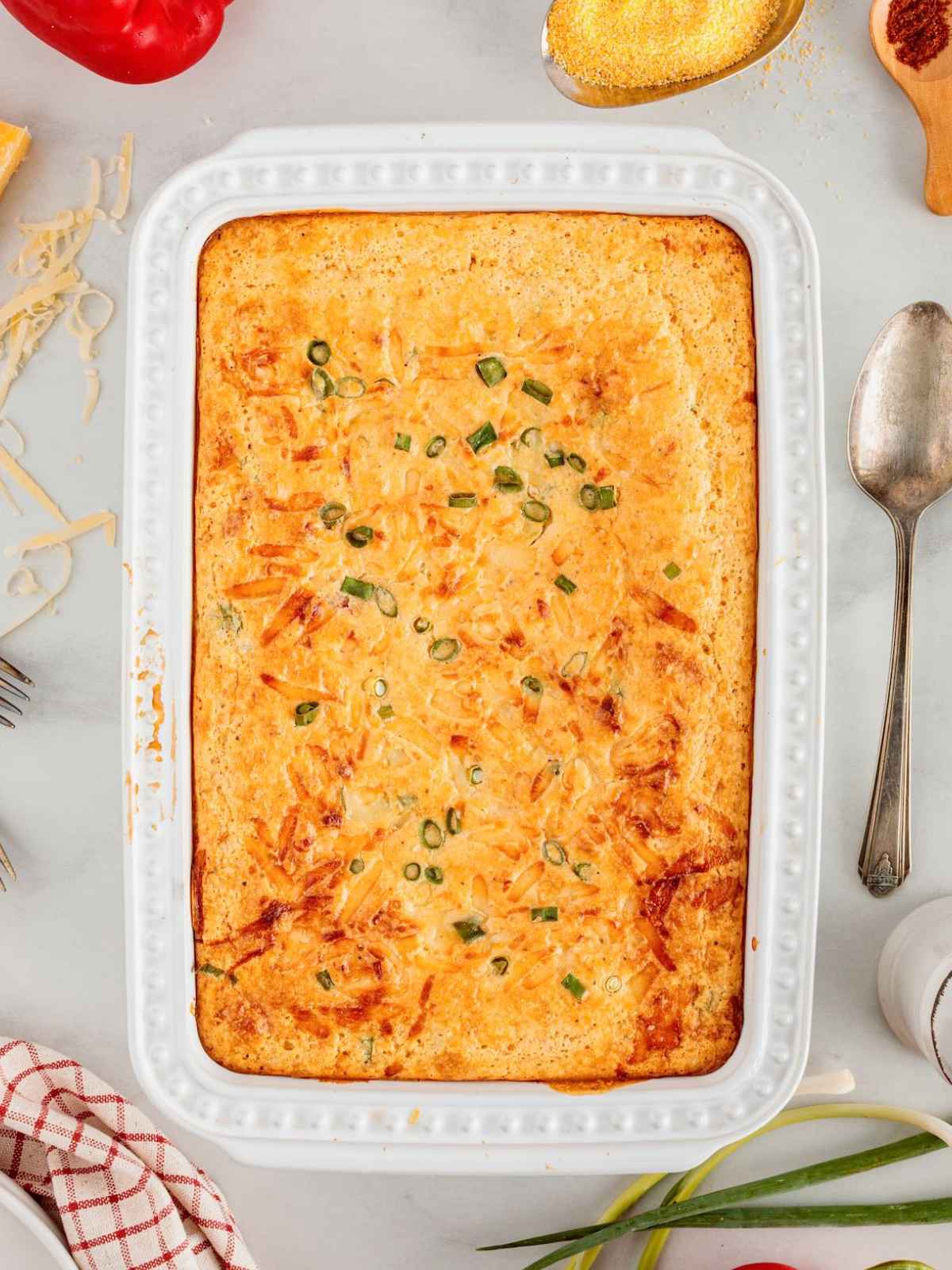 Baked corn casserole topped with shredded cheese and green onions in a white rectangular baking dish.