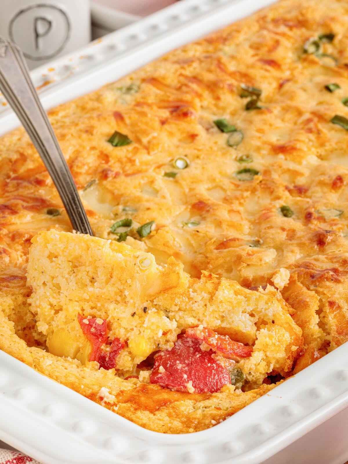 A serving of baked corn casserole with peppers and green onions being lifted from a white baking dish with a spoon.