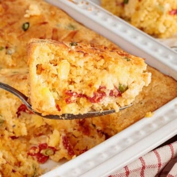 A spoonful of golden cornbread with corn kernels and red peppers, lifted from a baking dish.