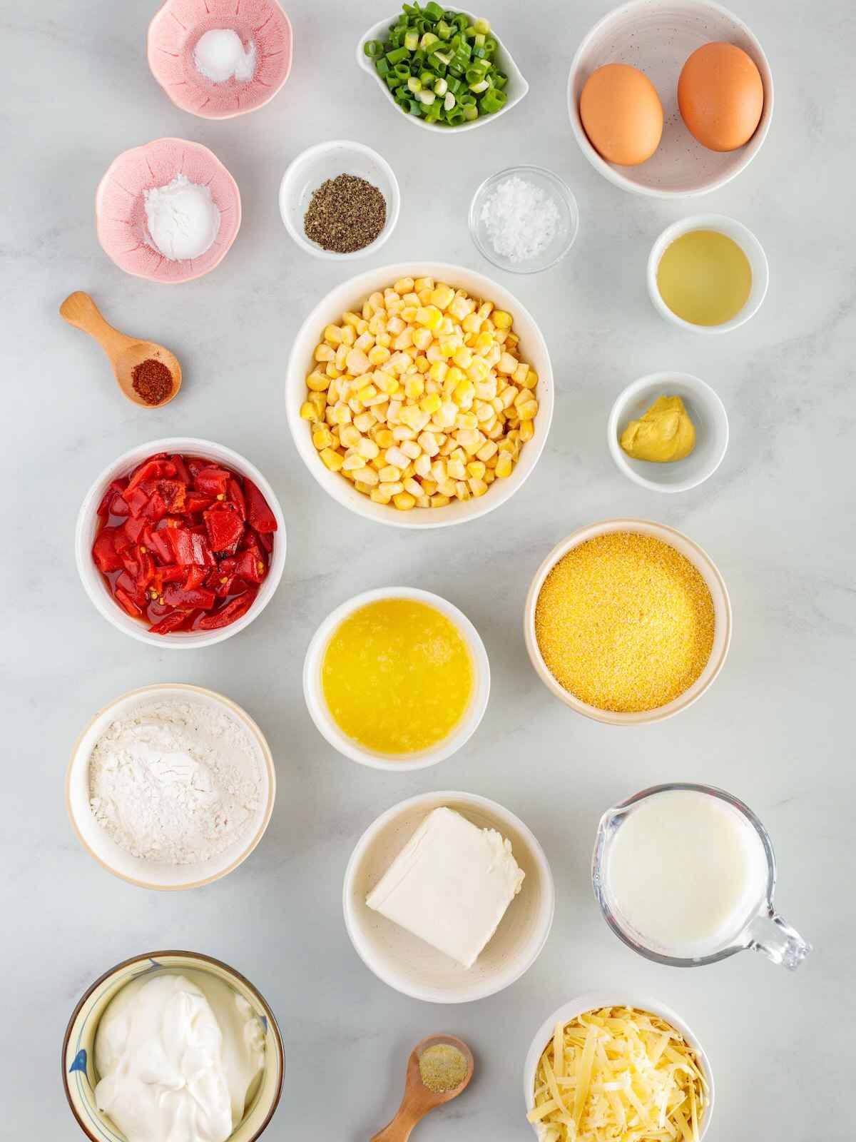 Top-down view of various ingredients in bowls, including corn, flour, cheese, eggs, and milk, arranged on a light surface.