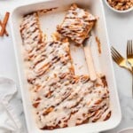 Iced pumpkin cinnamon roll cake in a white baking dish with a spatula, some pieces served, walnuts on the side.